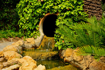 mini kaskada wodna w ogrodzie, woda płynąca z dzbana w korycie między kamieniami, mały strumień w ogrodzie, waterfall in garden © kateej