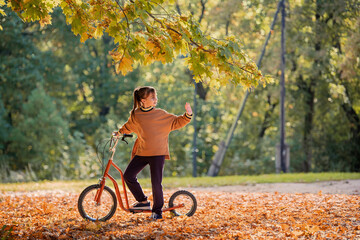 Little girl riding kick scooter through yellow leaves in autumn park.