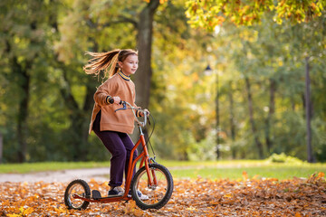 A young caucasian woman riding a scooter on a path covered with autumn leaves in a park during sunny day.