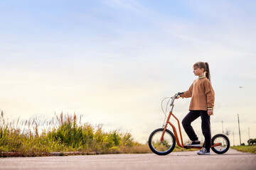 A young girl stands with her orange kick scooter on an empty road next to a grassy area