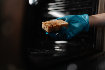 Cropped shot of cleaning company worker in blue rubber glove holding dirty sponge, scrubbing soiled gas oven covered in grease stains and splashes, tackling mess in kitchen environment, closeup.