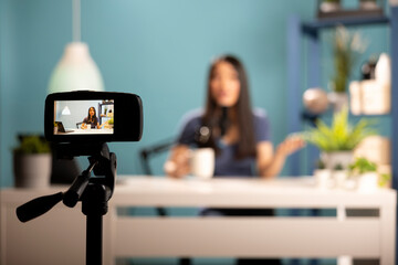 Asian woman blogger seated at desk in a professional studio setting, holding a cup of coffee and...