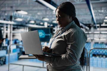 Data center female system administrator using laptop, configuring storage devices for minimal latency in AI workloads. African american woman using notebook in server farm to build neural networks