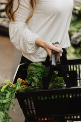Woman’s hands holding a black shopping basket filled with fresh organic lettuce and carrots during grocery shopping in a supermarket produce aisle