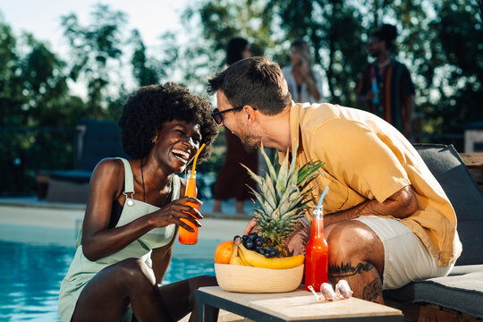 Happy multiethnic couple enjoying drinks and fruits by the pool during summer party - Powered by Adobe