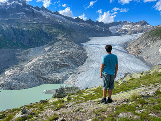 Naklejka premium Man stands admiring the stunning view of vast river of ice flowing into a turquoise glacial lake. Dramatic mountain peaks rise majestically above Rhone Glacier, accentuating the grandeur of landscape.