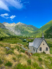 Obraz premium Charming stone chapel with a spire stands in a vibrant green valley, surrounded by towering, rugged mountains. Energetic dog runs through the grass during a scenic walk through Swiss alpine valley.