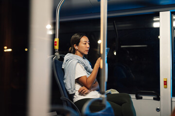 Young woman using smartphone and wearing earbuds while riding bus at night