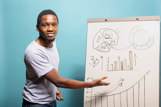 Young businessman in casual t shirt, gesturing to graphs on a flipchart, standing against an isolated blue background. Black male individual presenting business data charts on a whiteboard.