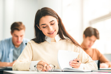 Fototapeta premium New Highschool Year On Campus. Positive Beautiful Young Adolescent Student Lady Sitting At Table In Modern Classroom, Doing Test Exercise, Reading Task, Enjoying Education And Learning