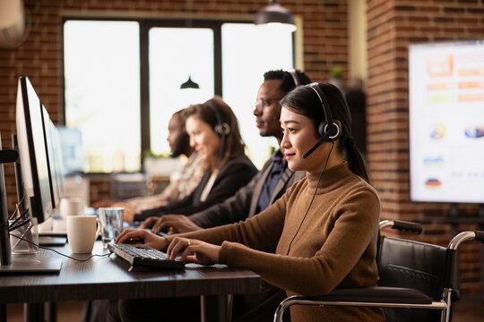Dedicated female professional in wheelchair, wearing wired headphones, focuses on tasks at her office desk. Asian woman entrepreneur engages with online customers, updating records on her computer.