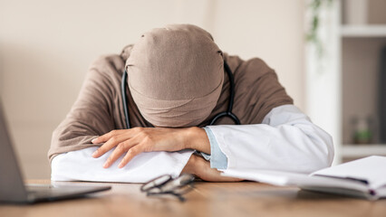 Overworked female doctor woman in hijab having nap at workdesk, working with laptop in clinic. Exhausted physician young lady leaning on her hands, needs for break during long working day