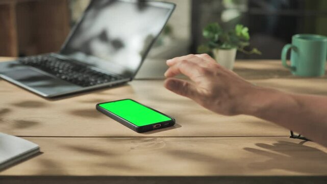 female hand tapping touching on smart phone mobile green screen on wooden table with laptop computer in the background