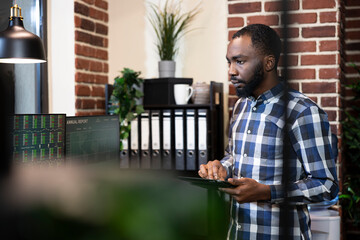 African american man with tablet, working night shift, examining financial reports on computer...