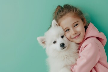 Smiling little girl in pink hoodie hugging fluffy white spitz dog on pastel green background in close portrait