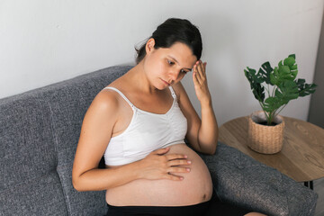 Pregnant woman sitting on grey couch in white top with hand on forehead and bare belly exposed, expressing discomfort or fatigue, indoor scene with potted plant, maternity concept