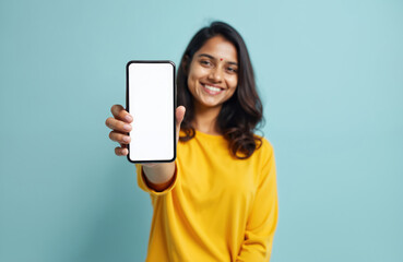 Fototapeta premium Young Indian woman presents blank smartphone screen. Happy girl shows device with empty display. Modern tech, app, design, advertising concept. Female holds mobile phone on blue background. Smiling