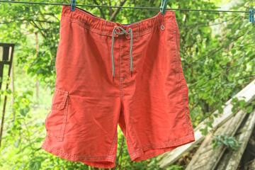 one wet shorts made of red fabric hanging on a wire against the background of green vegetation on a summer street