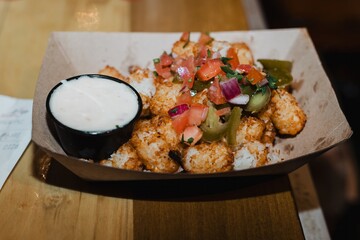 Paper tray of loaded tater tots topped with pico de gallo and pickled jalapeños alongside a cup of...