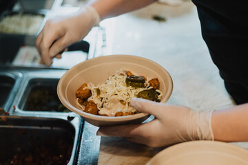 Gloved hands holding a bowl of loaded tater tots topped with shredded cheese, pickled jalapeños and sauce in a kitchen prep line