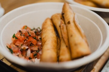 Compostable bowl of crispy fried taquitos served with pico de gallo on a wooden counter in a restaurant kitchen