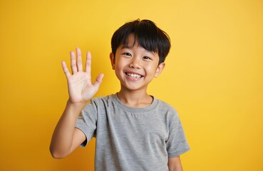 Portrait of smiling Asian boy in grey casual t-shirt waving hand against vibrant yellow background. Happy child showing palm, greeting, saying goodbye. Cheerful student at studio.