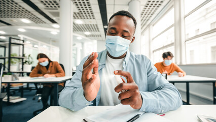 Hygiene And Protection Concept. African american guy applying antibacterial sanitizer spray on hands, wearing surgical face mask, sitting in classroom. Prevent the spread of infection and germs