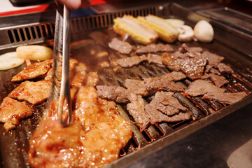 A person uses metal tongs to cook marinated pork and sliced beef on a hot tabletop grill during a Korean BBQ or Japanese Yakiniku meal. 