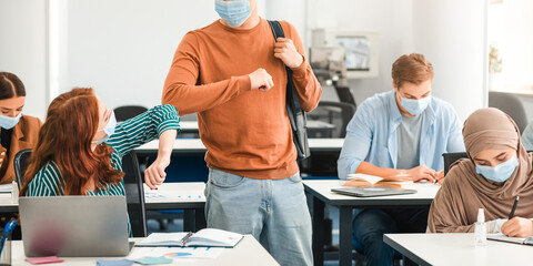 Elbow Bump. Diverse multicultural people wearing protective surgical face masks greeting each other at classroom. Asian guys standing with backpack, woman sitting at table, students avoid touching
