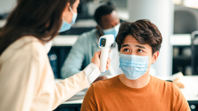 Teacher using digital medical contactless infrared electronic thermometer, measuring and checking body temperature, screening asian student in protective mask, preventing contagious diseases spread