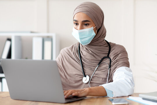 Young female doctor in headscarf and protective face mask using laptop while working at clinic, typing patient data or prescription, physician working during covid-19 pandemic at hospital, free space