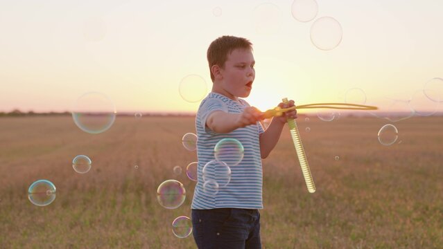 Child boy blows soap bubbles, having fun playing on field in sunset. Child boy plays with soap bubbles in park in sun nature. Happy childhood concept. Cheerful kid smiling, holiday in summer outdoors
