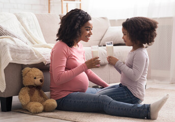 Healthy Drink. Little afro girl and her pregnant mother drinking milk sitting on floor at home,...