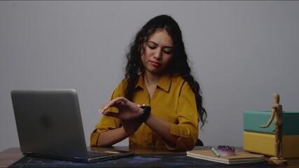 South Asian woman working on her laptop and checking her watch, managing time during a busy day - Powered by Adobe