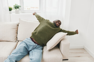Obraz premium Happy black man relaxing on a couch in a cozy living room, wearing a green sweater and jeans, with an expression of joy and comfort