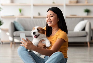 Attractive young asian woman holding fluffy puppy and taking selfie on smartphone while enjoying time together at home, happy jack russel terrier owner taking photos with dog, copy space