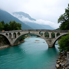 Fototapeta premium Elegant stone bridge with arches over a turquoise river, set against majestic misty mountains and an overcast sky.