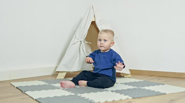 Cheerful toddler boy playing at home on a rug and sitting in a wigwam. A house for a child in an apartment. A wigwam for a child. A child in the living room is mischievous and laughing