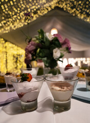 Catering service for a birthday. Closeup view of the desserts table with modern tiramisu and cheesecake glass cups with a beautiful floral arrangement. The garden lights and tent in the background