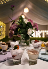 Catering service for a birthday. Closeup view of the desserts table with modern tiramisu and cheesecake glass cups with a beautiful floral arrangement. The garden lights and tent in the background	
