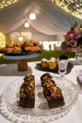 Sweet desserts table in a birthday party. Closeup view of dark chocolate brownies with nuts in a glass dish. The white tent, lights and floral arrangement in the background