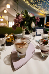Catering service for a birthday. Closeup view of the desserts table with modern tiramisu and cheesecake glass cups with a beautiful floral arrangement. The garden lights and tent in the background	
