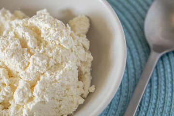 Close-up of fresh farmers cheese in a ceramic plate with a metal spoon, styled on a textured light blue placemat. A rustic and healthy food concept with natural tones and light