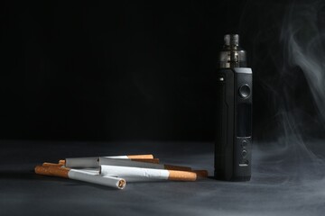 Electronic smoking device and cigarettes in smoke on dark table against black background, closeup