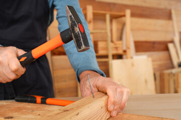 Mature carpenter hammering nail into wooden plank at table in shop, closeup