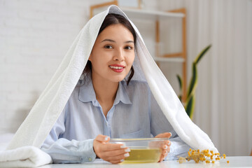 Young Asian woman with towel doing steam inhalation at table in bedroom