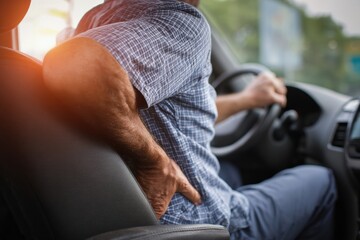 Mature man driving a car, experiencing back pain, touching his hip while keeping one hand on the steering wheel during a journey
