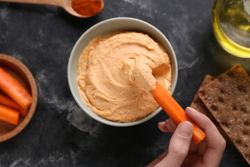 Woman eating tasty hummus with fresh carrot sticks on black grunge background