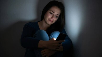 Woman sitting in dark corner crying with tears streaming down face while holding phone