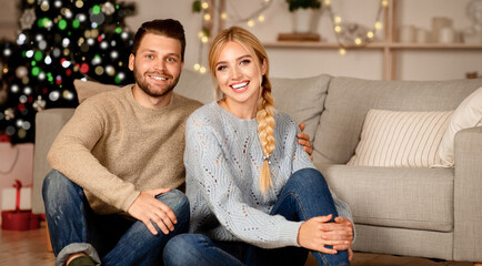 Beautiful young couple celebrating Christmas together at home, sittng on the floor by Christmas tree and embracing, copy space. Happy man and woman posing on camera on xmas eve, family portrait